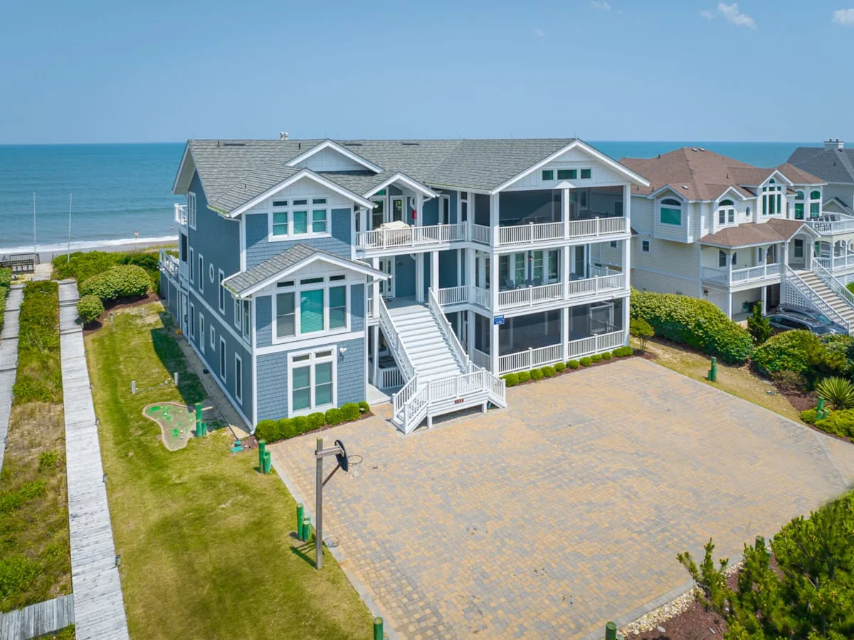 A large coastal house stands beside a paved area, with the ocean visible behind it.