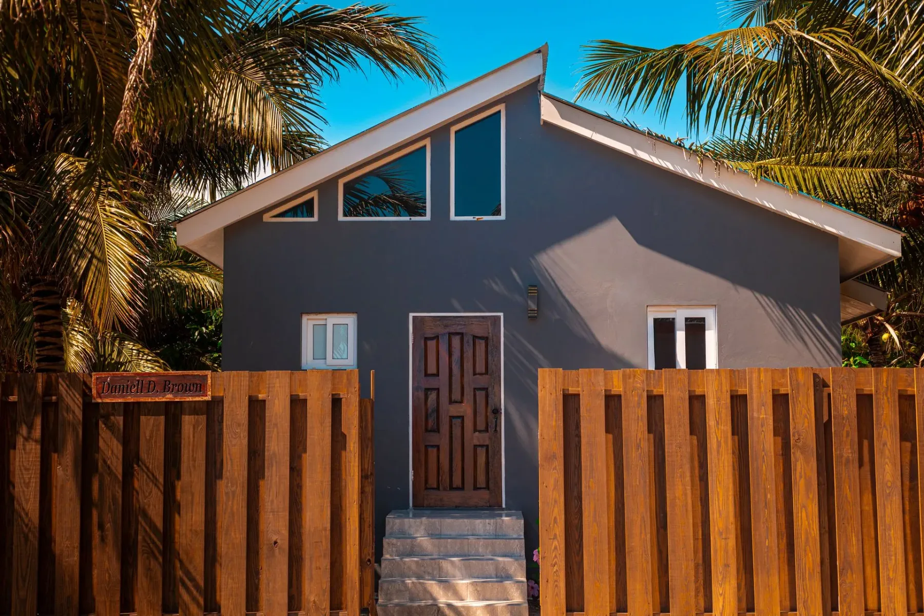 Gray house behind a wooden fence, with palm trees.