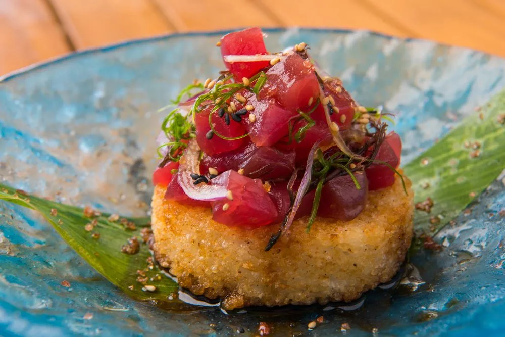 Un aperitivo emplatado con ingredientes rojos picados y hierbas sobre un plato azul.