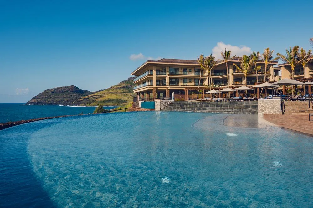 Un edificio junto a una piscina amplia, con palmeras y asientos, da al mar.
