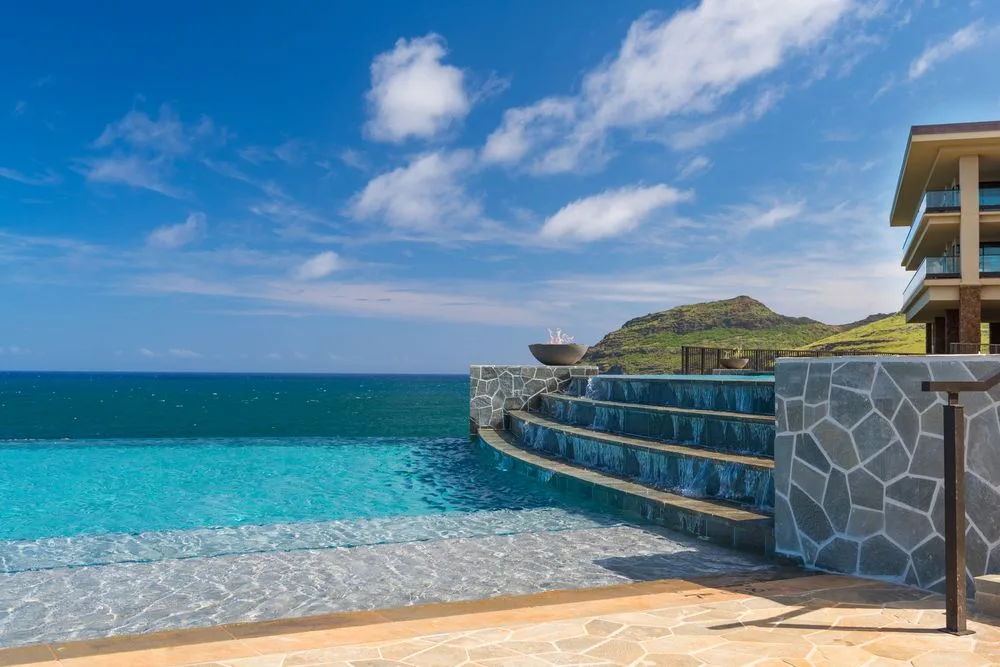 Una piscina al aire libre mira al mar, con una terraza de piedra y colinas verdes al fondo.