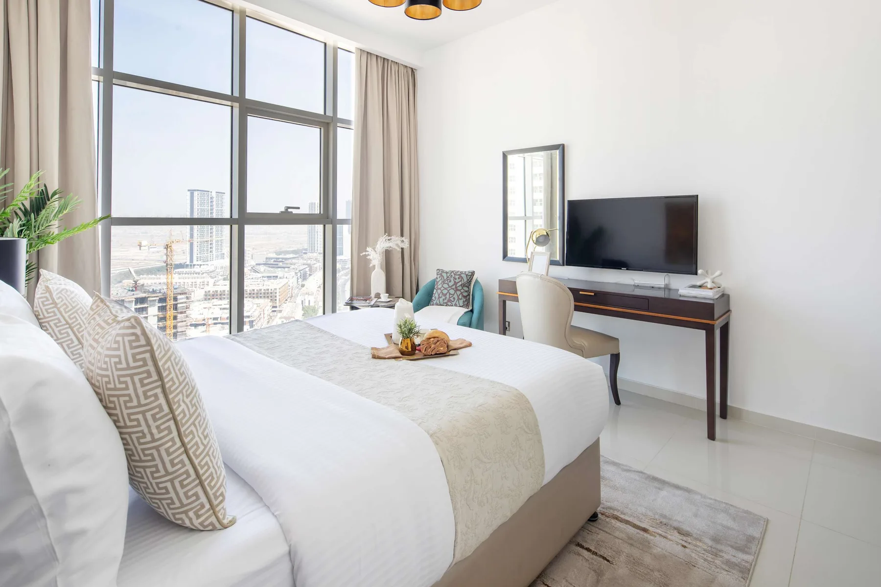 Un dormitorio luminoso con una cama bien tendida junto a grandes ventanas y un televisor.
