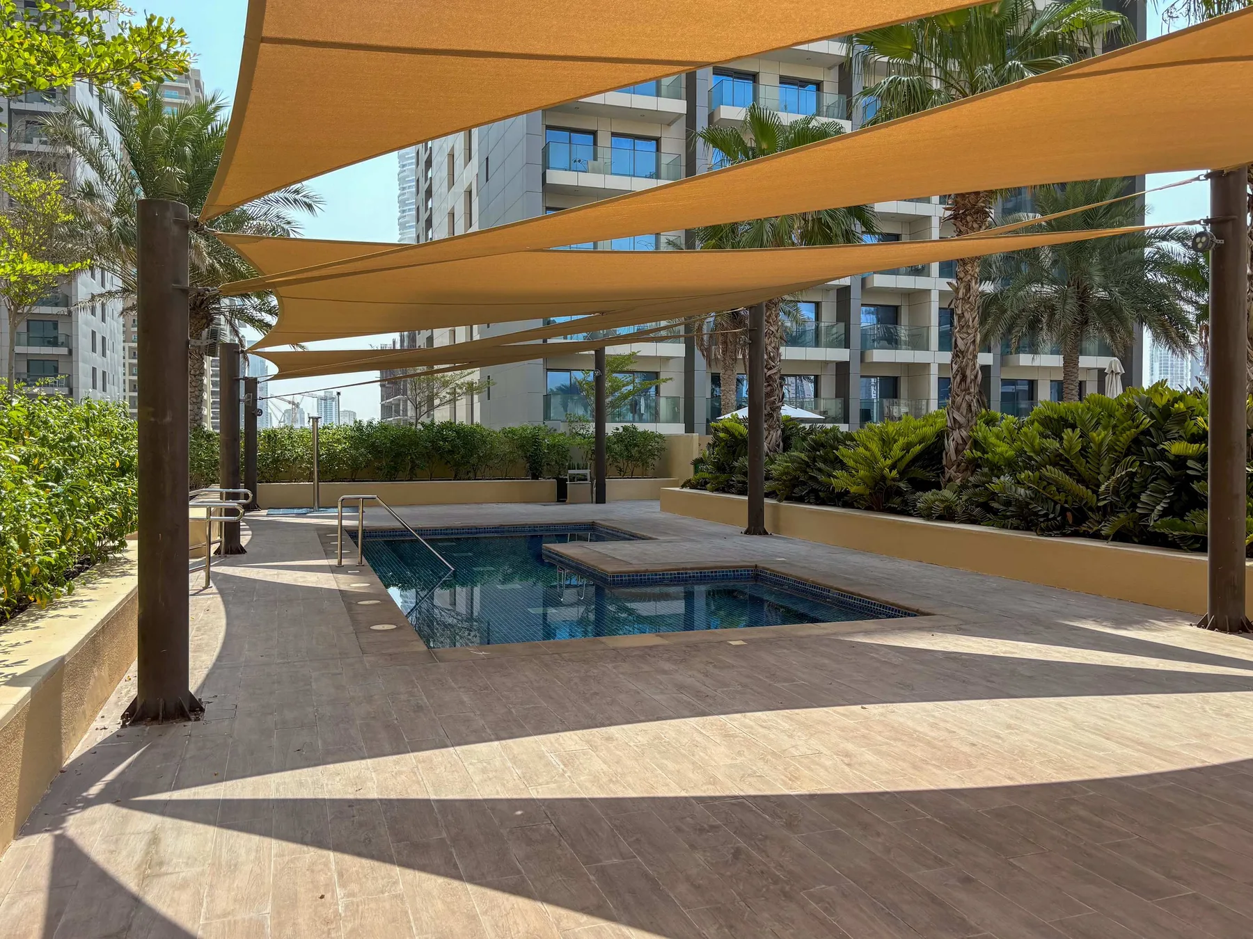 Una piscina al aire libre está bajo amplias velas de sombra, con vegetación y edificios de apartamentos cerca.