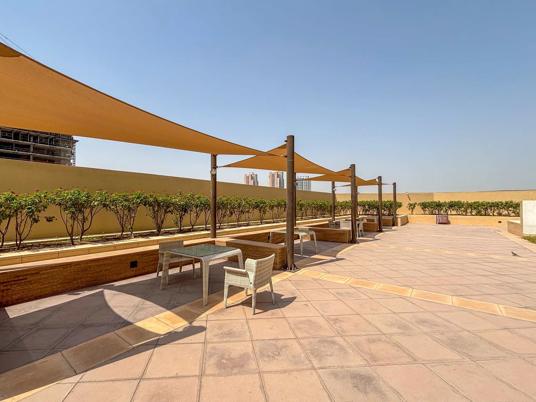 Una terraza exterior pavimentada tiene toldos de sombra y asientos junto a una hilera de arbustos.