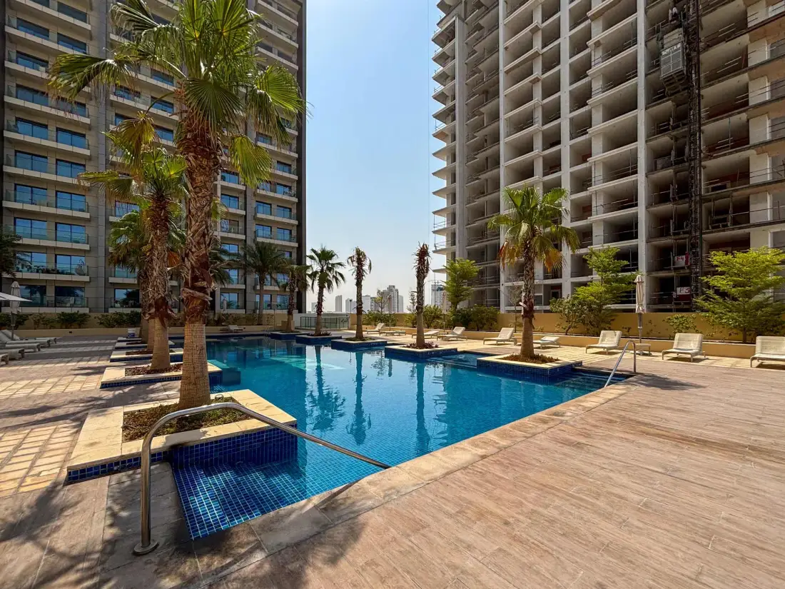 Una piscina al aire libre está entre dos edificios altos, con palmeras y tumbonas alrededor.