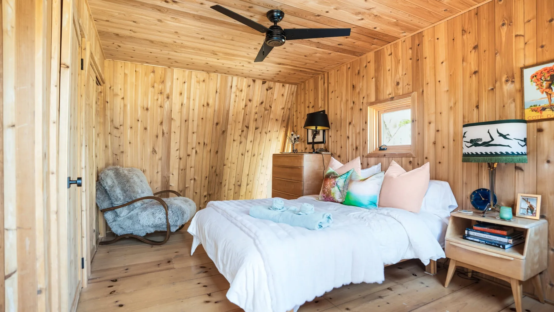 Un dormitorio revestido de madera con una cama, una cómoda pequeña y un ventilador de techo.