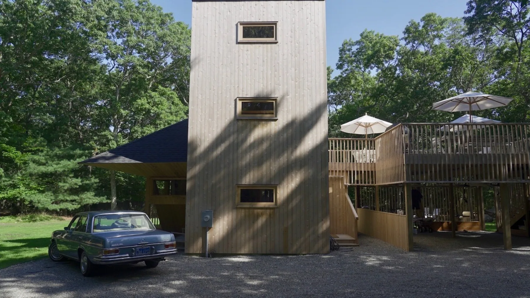 Un edificio revestido de madera está junto a una terraza elevada, con un automóvil estacionado delante.