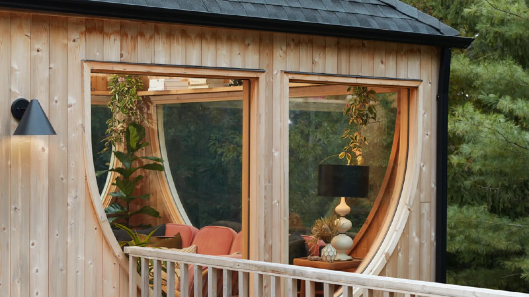 Una cabaña de jardín de madera con grandes ventanas curvas y plantas en el interior.