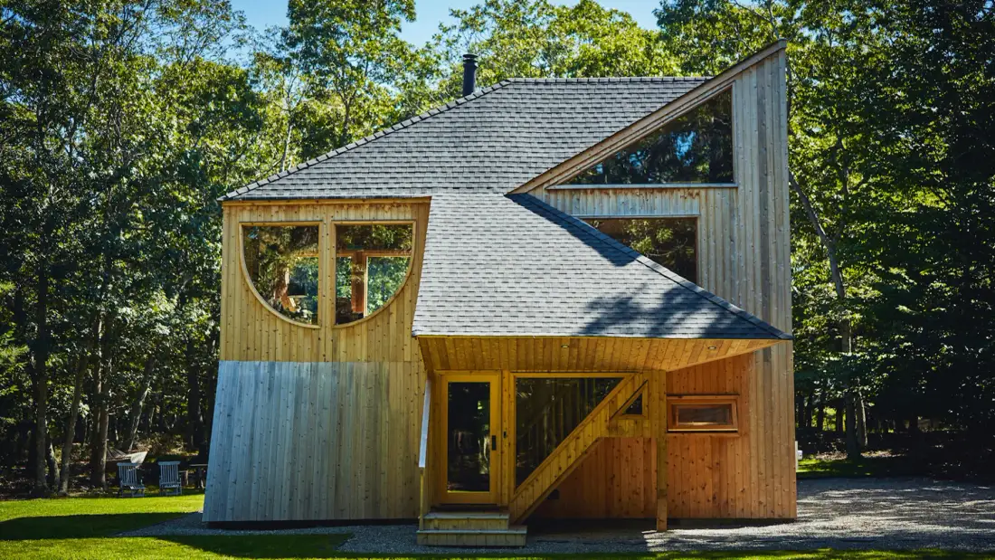 Una cabaña de madera entre árboles, con techo inclinado y ventanas grandes.