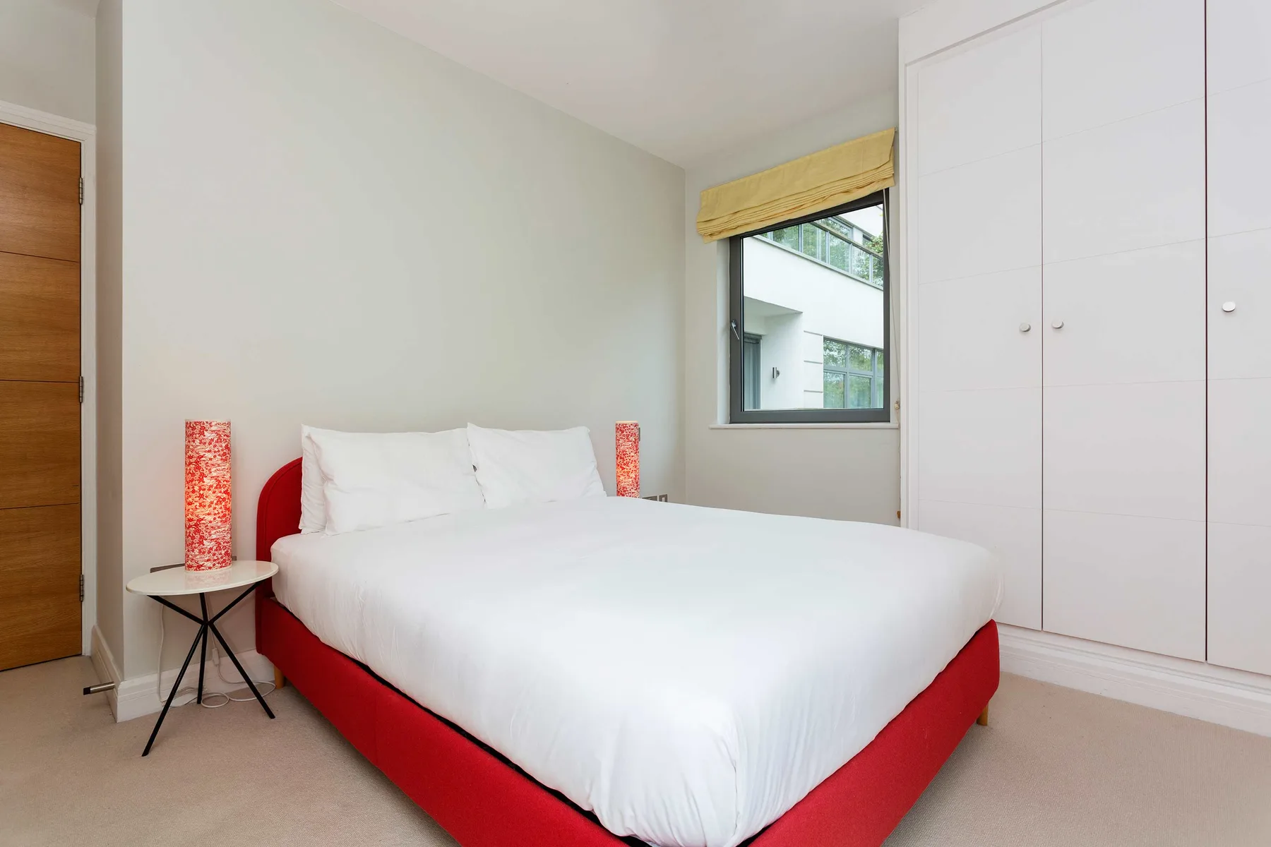 A bedroom with a white bed, red bed base, and built-in wardrobes.
