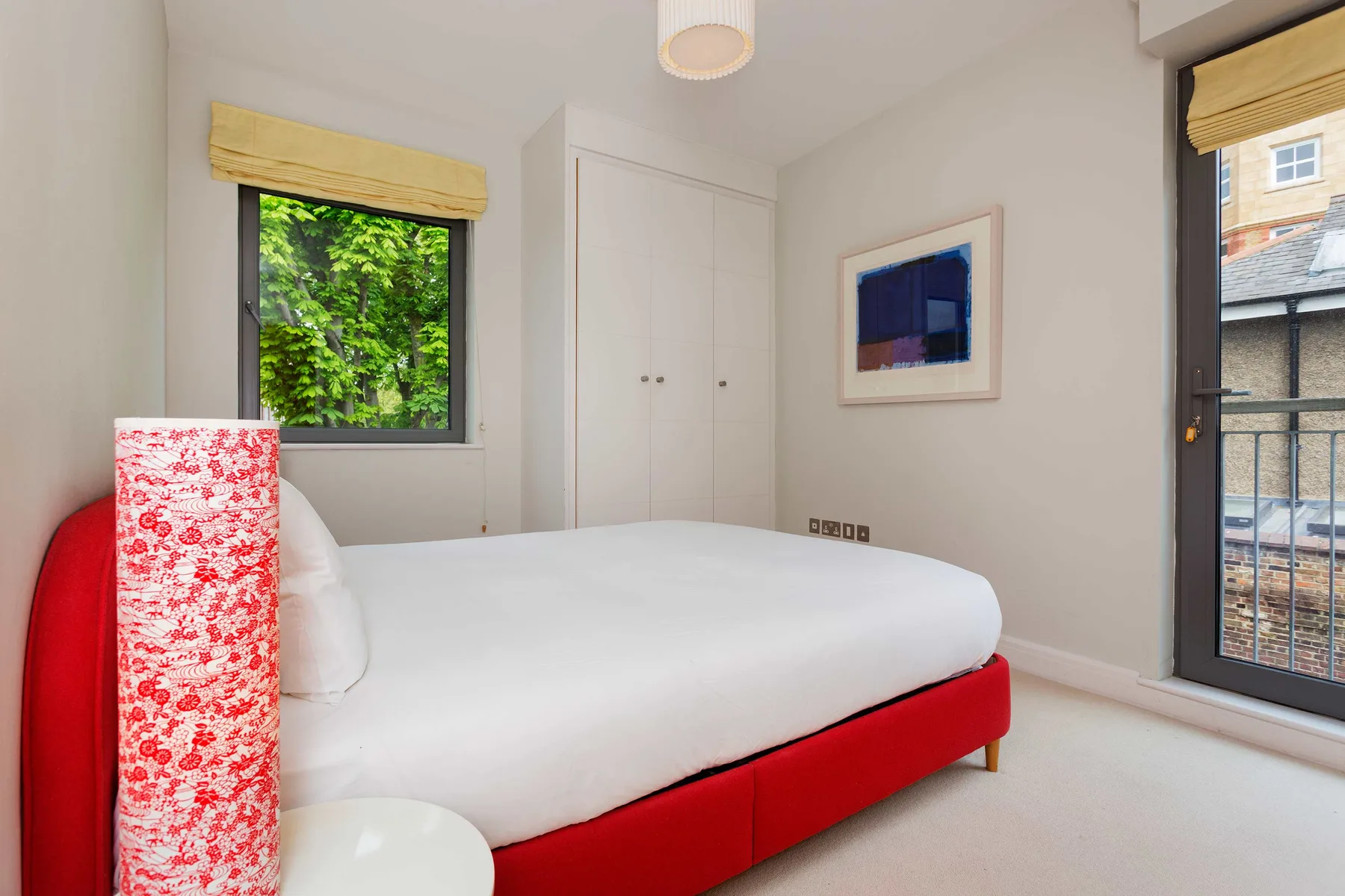 A bedroom with a white bed, red frame, and windows overlooking greenery and nearby buildings.