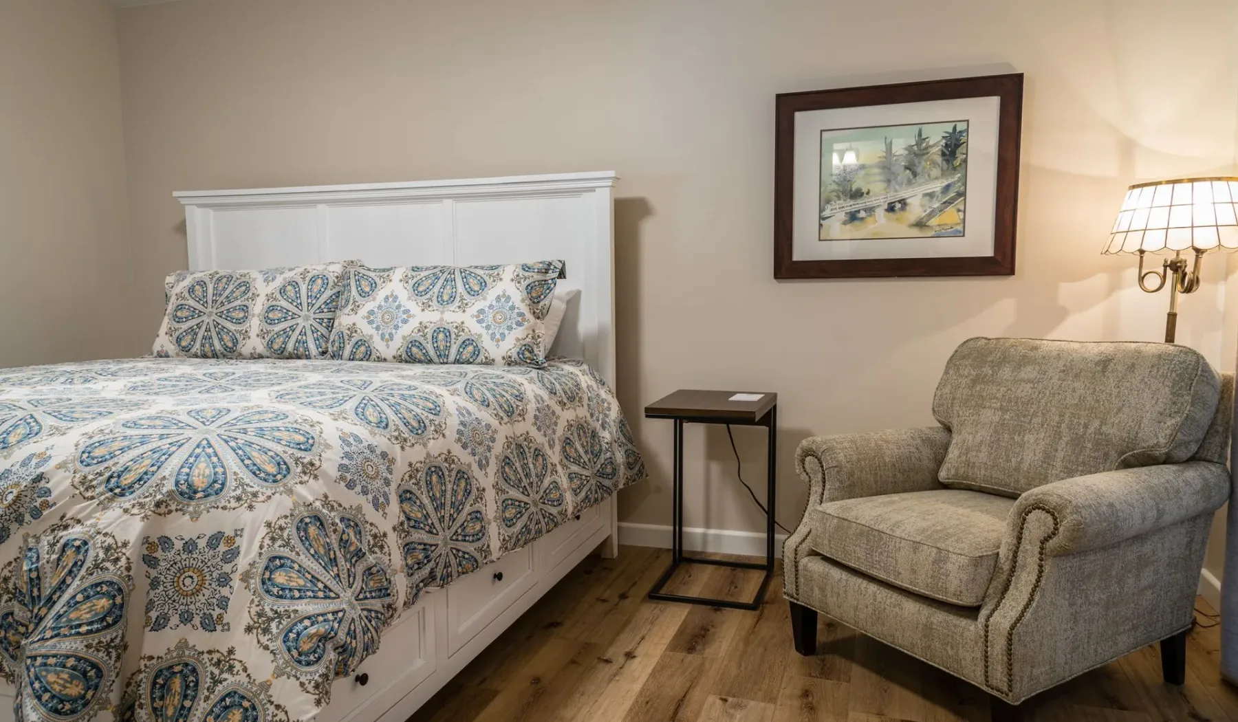 Un dormitorio con una cama estampada, un sillón y una mesita auxiliar.
