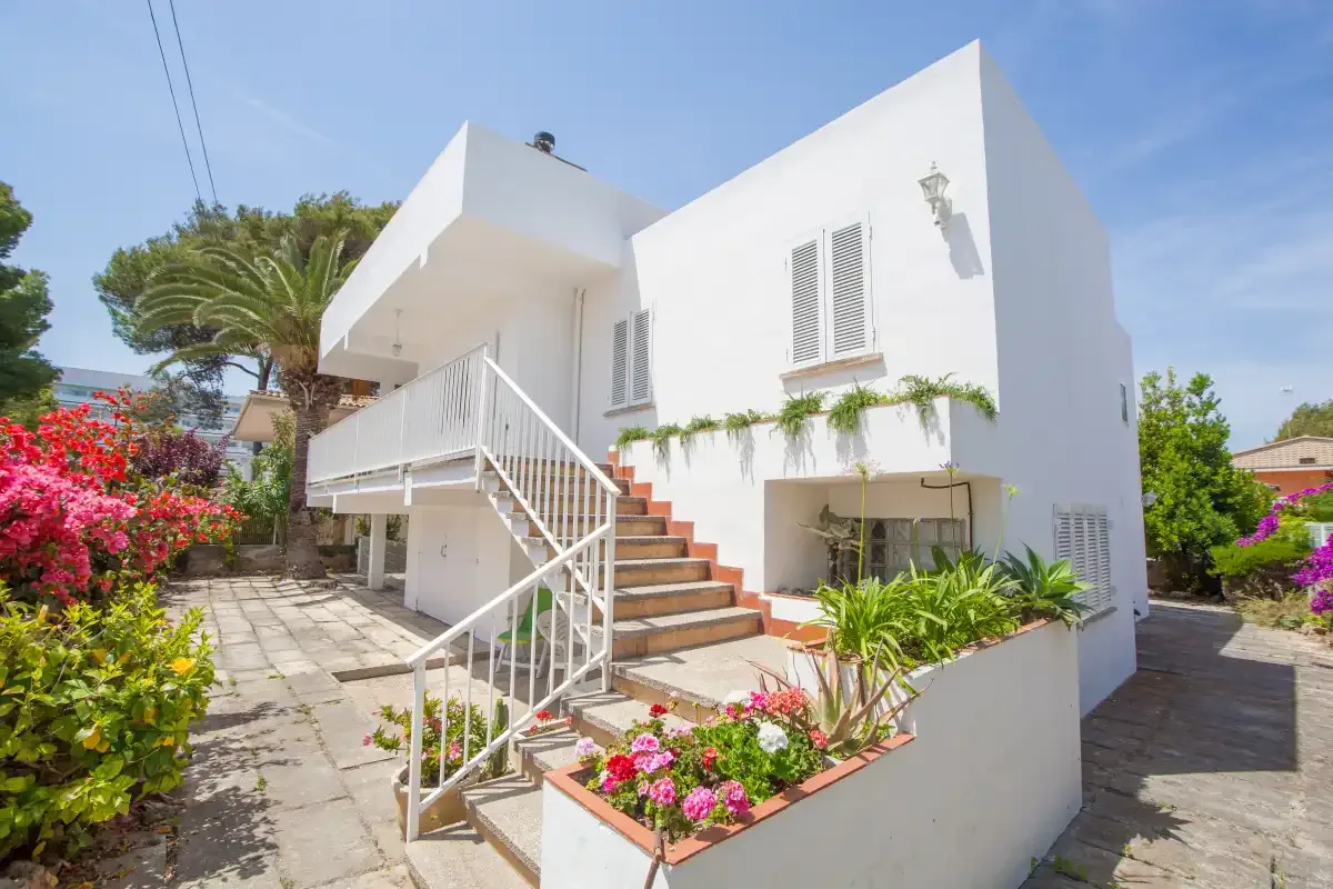 A white two-story house with an exterior staircase and flowering garden.