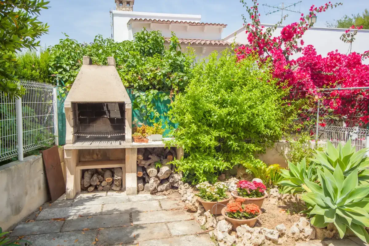 A garden patio with a stone barbecue, potted flowers, and lush greenery.