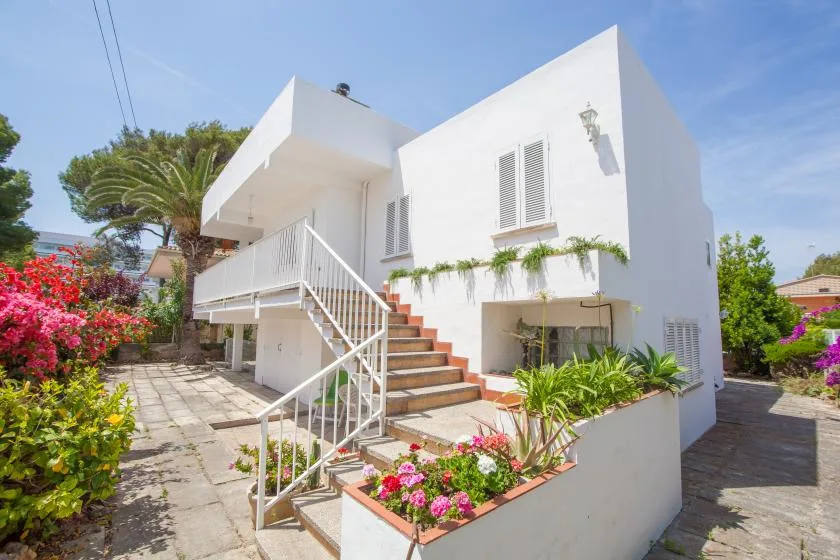 A white two-story house with exterior stairs, a patio, and flowering garden beds.