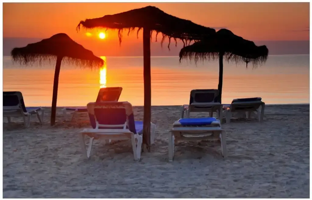 A beach at sunset with lounge chairs beneath thatched parasols.