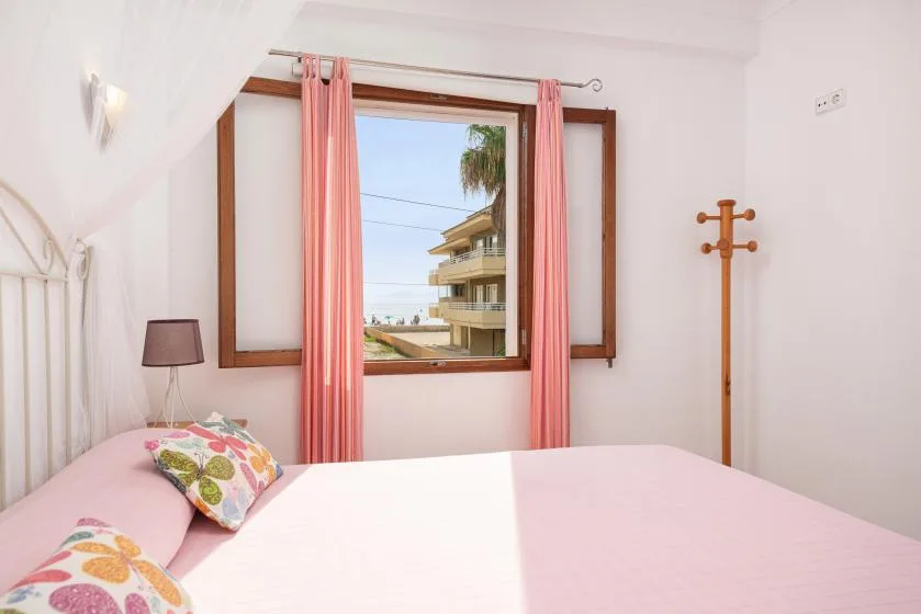 A bedroom with pink bedding and a window overlooking nearby buildings.