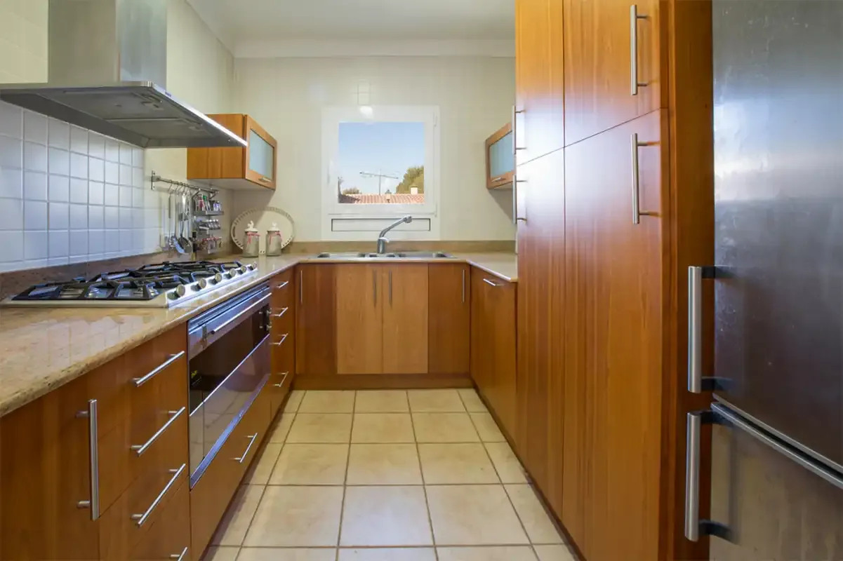 A narrow kitchen with wooden cabinets, tiled flooring, and a window above the sink.