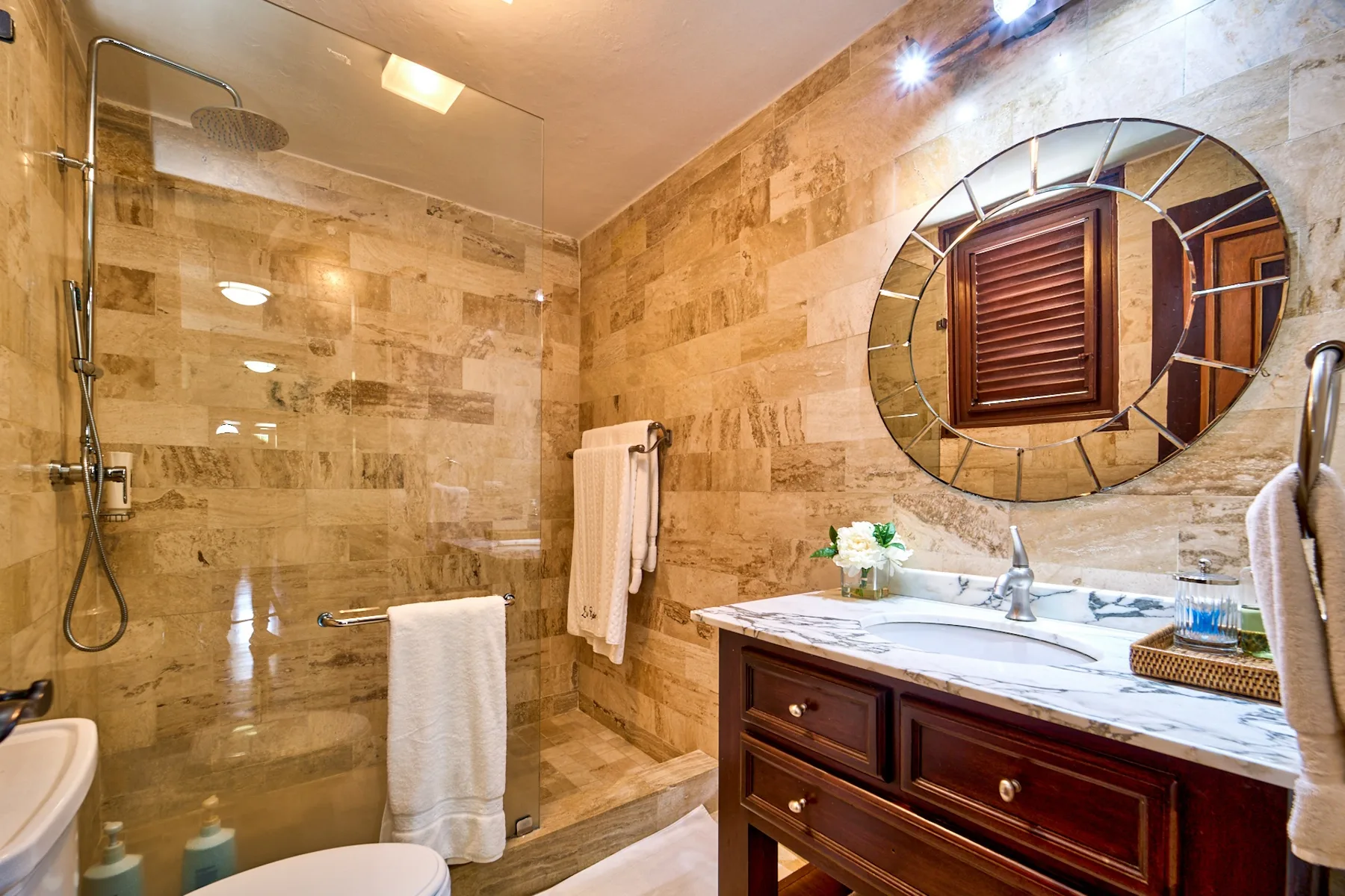 A tiled bathroom with a glass shower and a vanity beneath a round mirror.