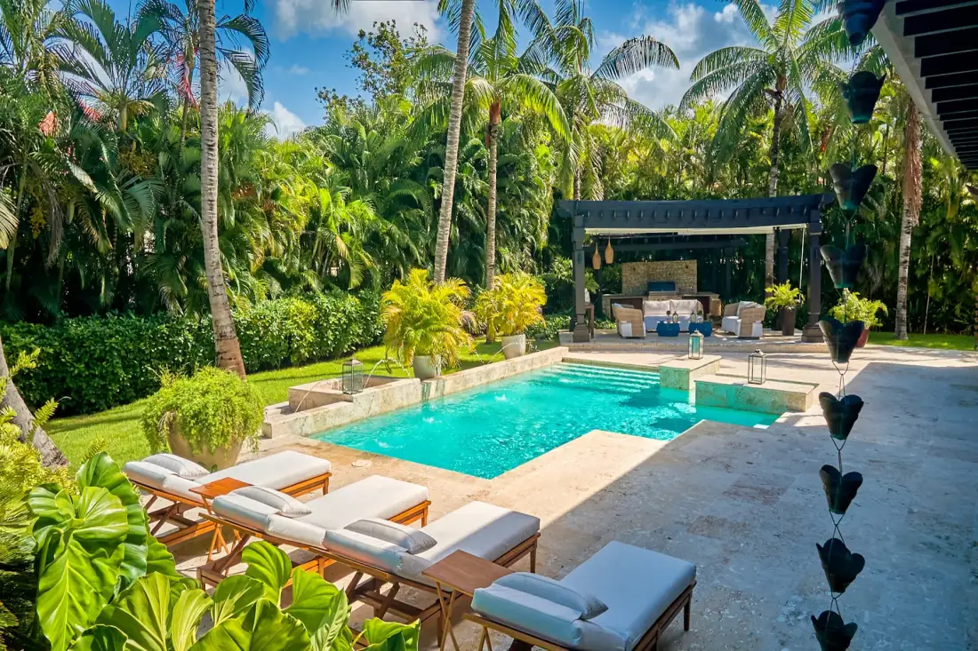 A swimming pool sits beside a paved patio surrounded by tropical plants.