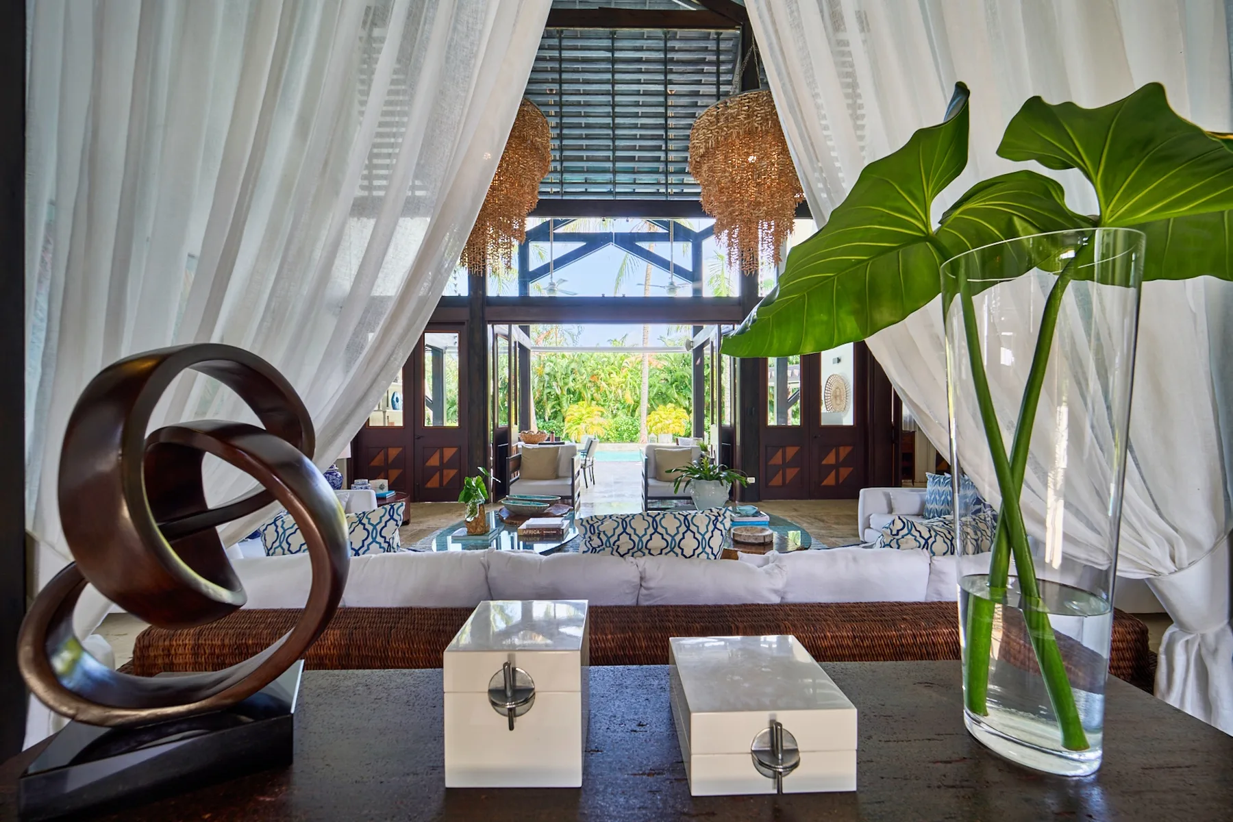 A bright living room with white curtains, a long sofa, and large leafy plants.