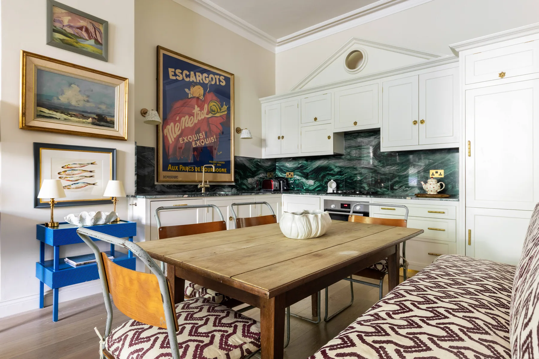 Una cocina y comedor luminosos con gabinetes blancos, una mesa de madera y asientos estampados.