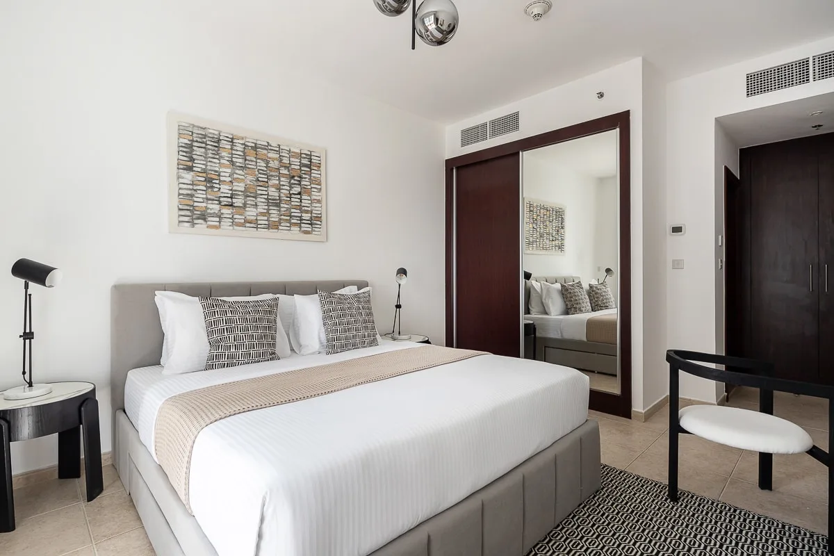 A bright bedroom with a neatly made bed, bedside tables, and a mirrored wardrobe.