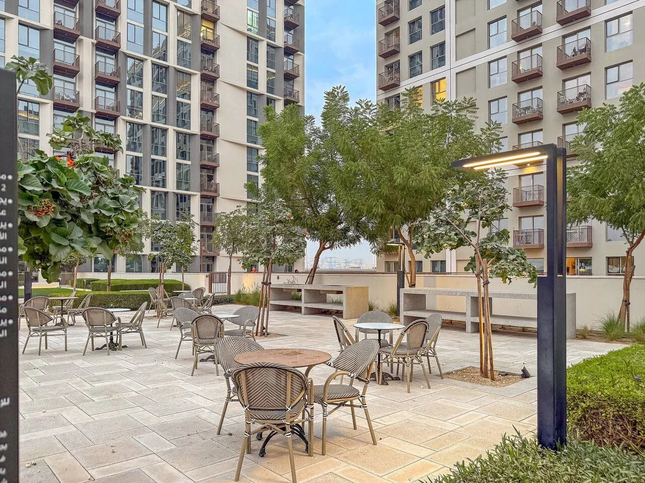 An outdoor courtyard with tables and seating beneath trees between apartment buildings.
