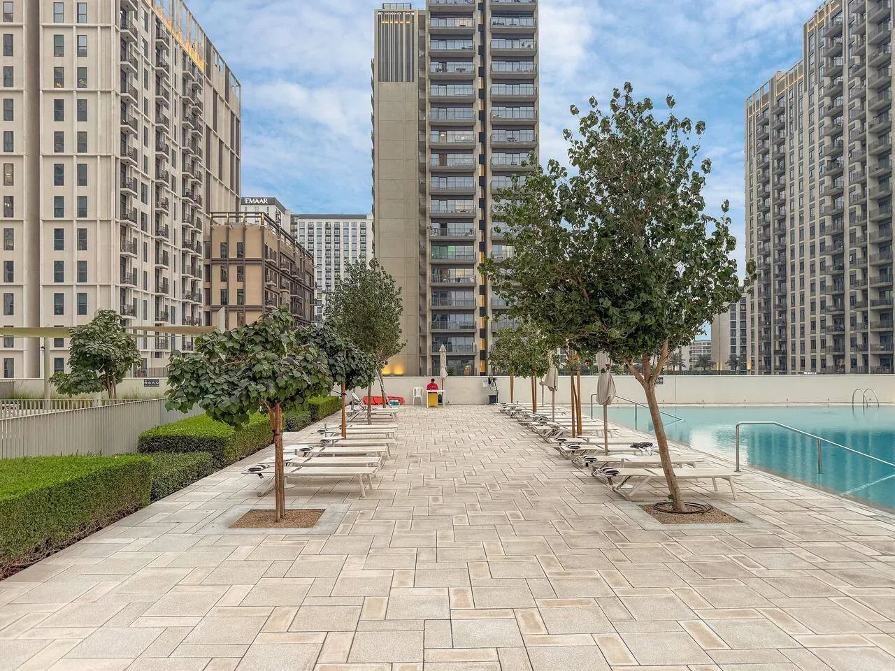 A paved poolside walkway runs between trees, loungers, and tall apartment buildings.