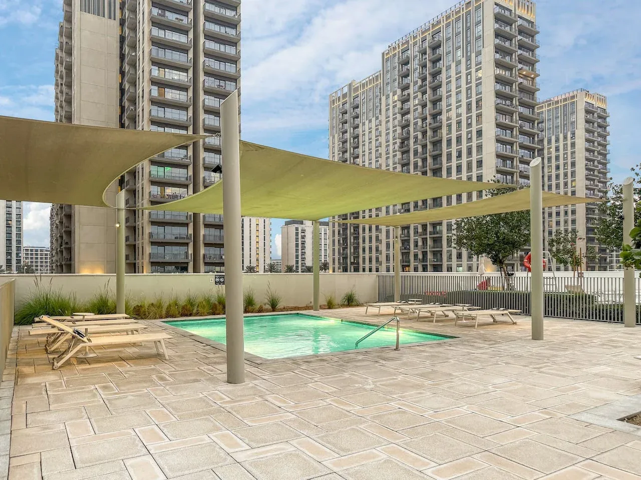 An outdoor pool and paved deck sit beneath shade sails, surrounded by tall buildings.