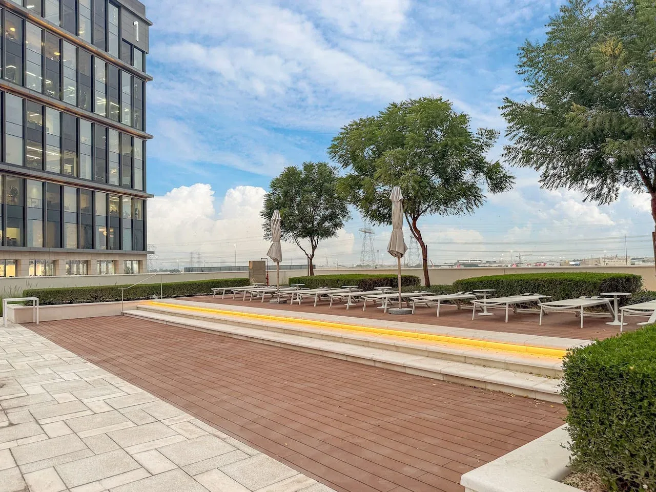 An outdoor terrace with lounge seating, closed umbrellas, and trees beside a modern building.