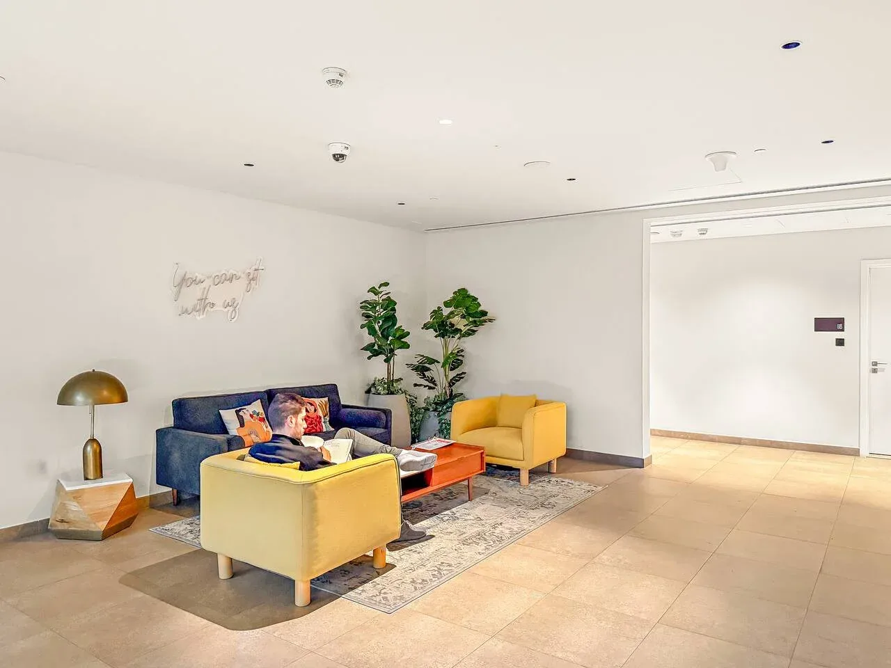 A bright lounge has colorful seating around a low table, with plants behind it.
