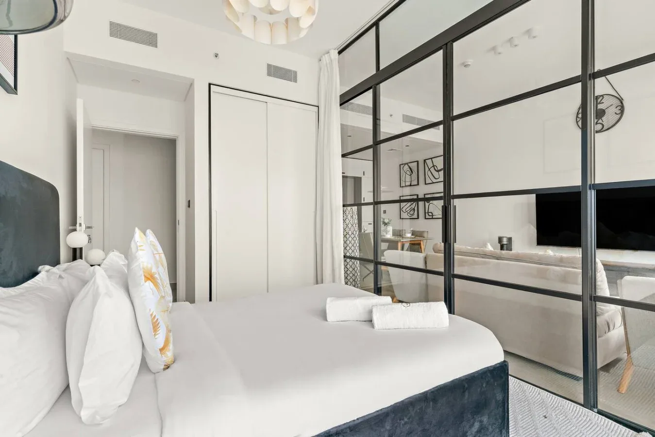 A bright bedroom with a neatly made bed beside a black-framed glass partition.