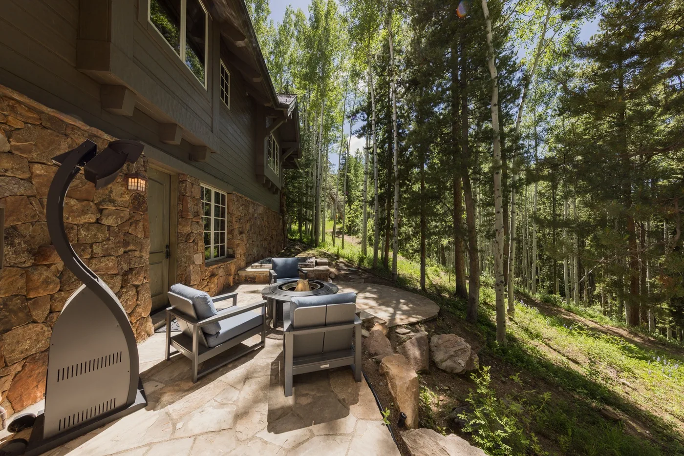 A stone patio beside a wooded home features outdoor seating and a fire pit.