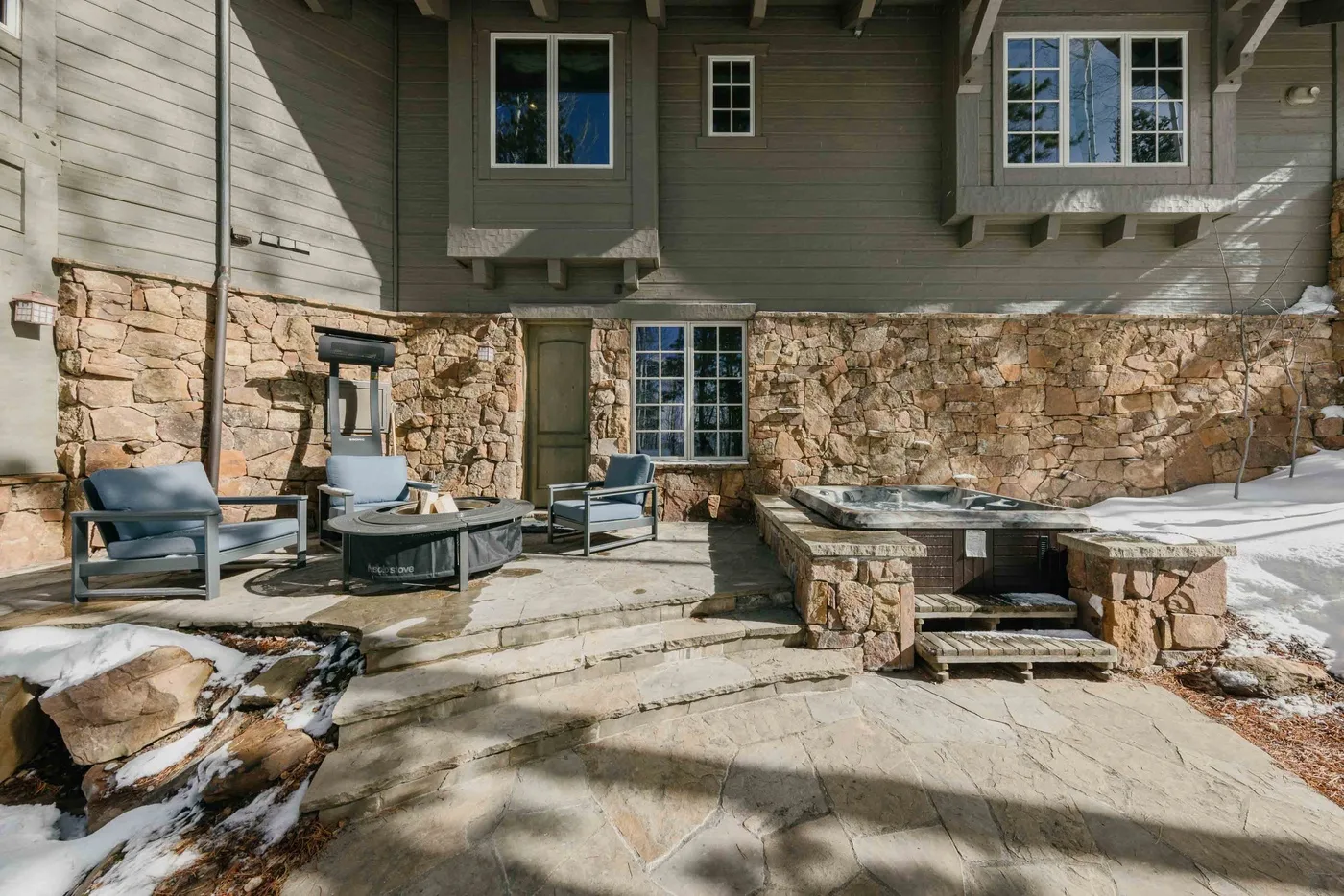 A stone patio with outdoor seating and a built-in fire pit beside a timber-clad house.