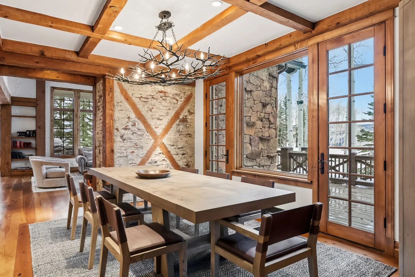 A rustic dining room with a long wooden table beside large windows.