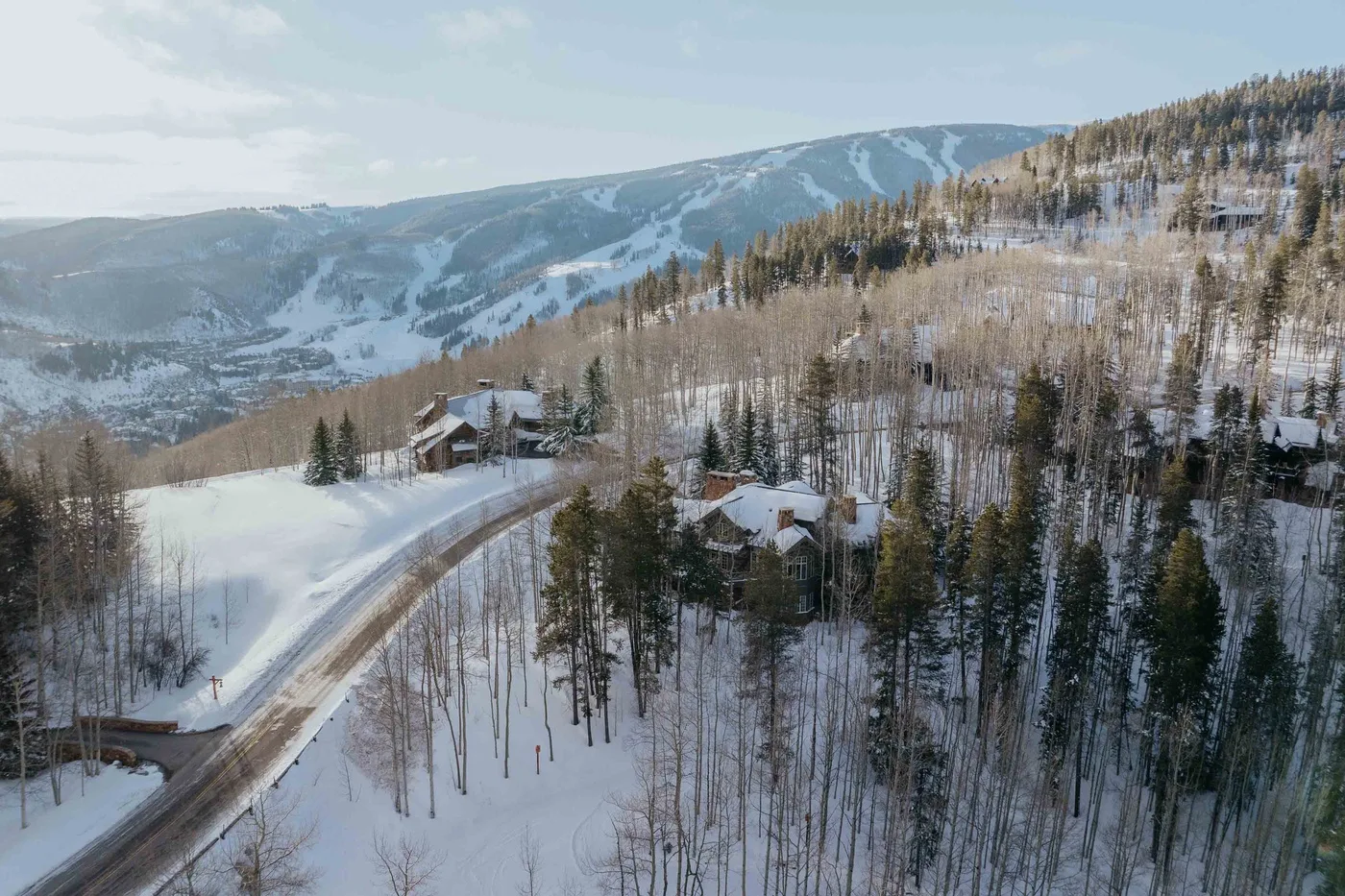 Aerial view of snow-covered cabins among trees, with mountain ski trails in the distance.