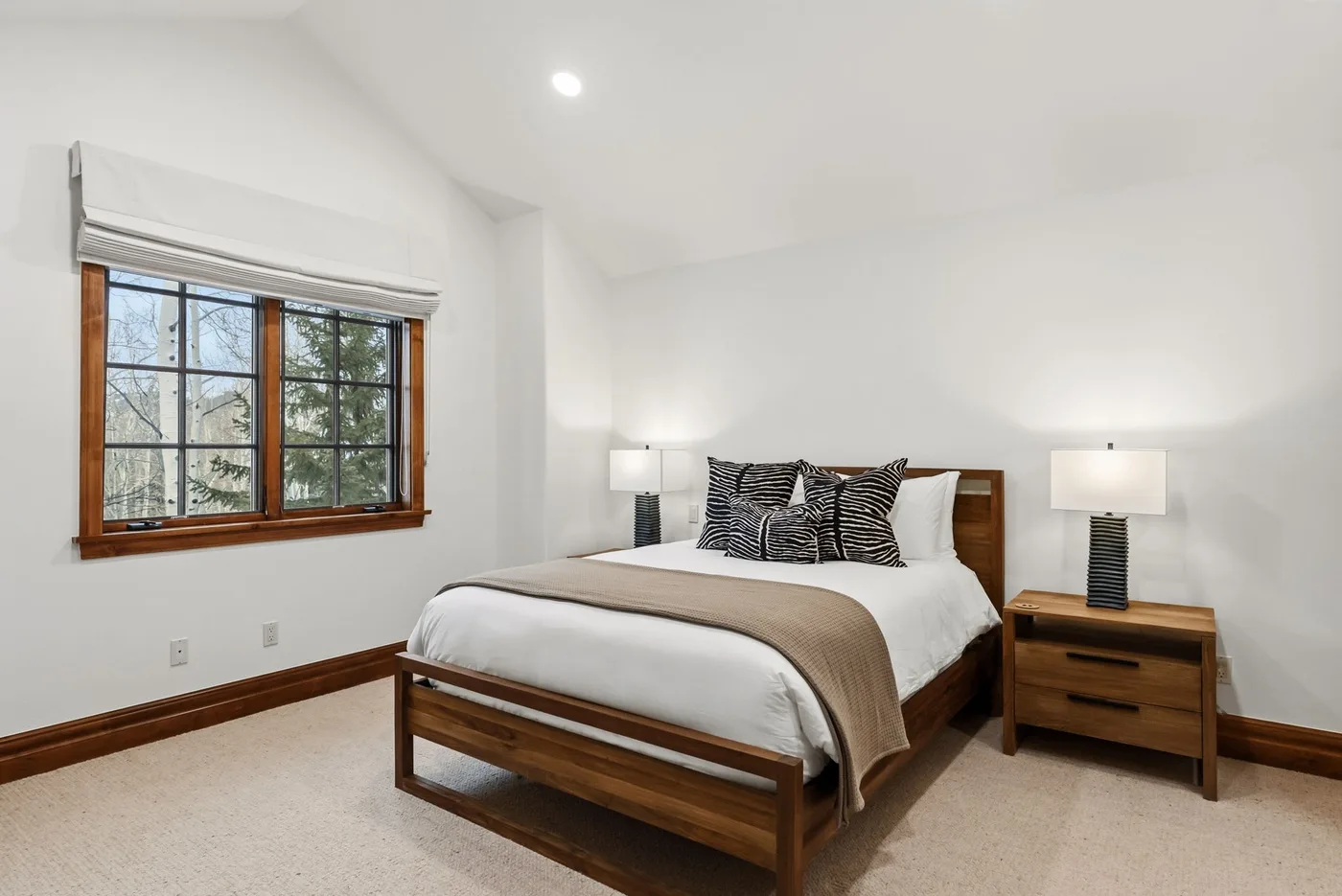 A bright bedroom with a wooden bed, neutral bedding, and a large window.