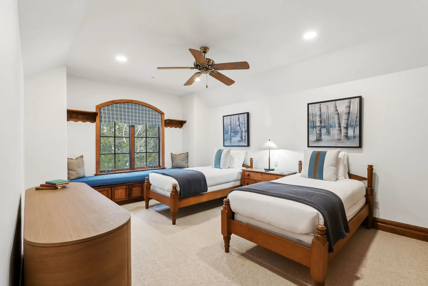 A bright bedroom with two beds, a large window, and a ceiling fan.