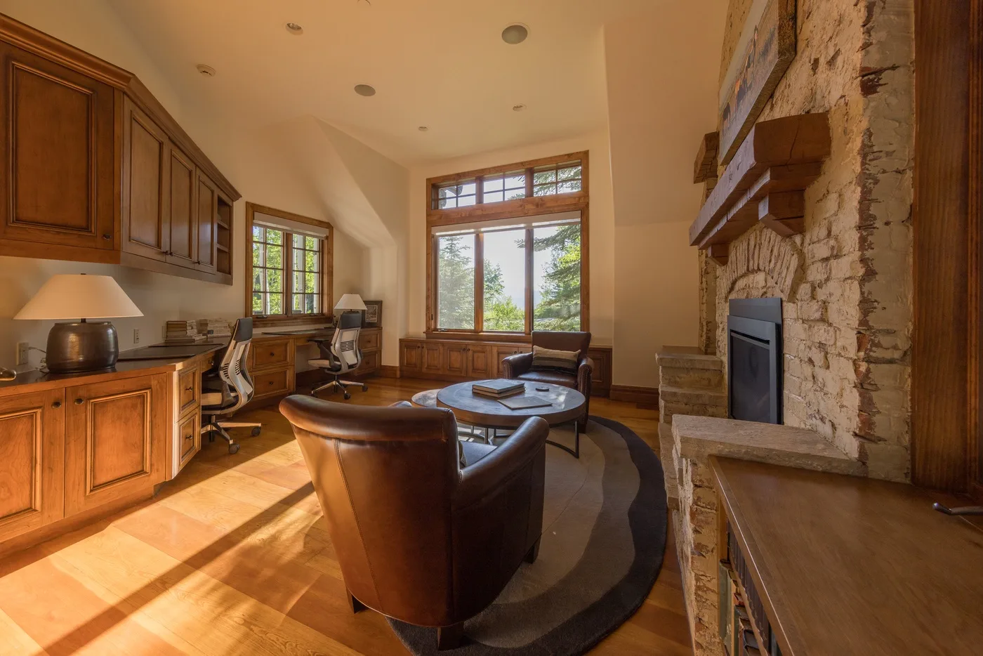 A warm living room with a seating area beside a stone fireplace and tall windows.