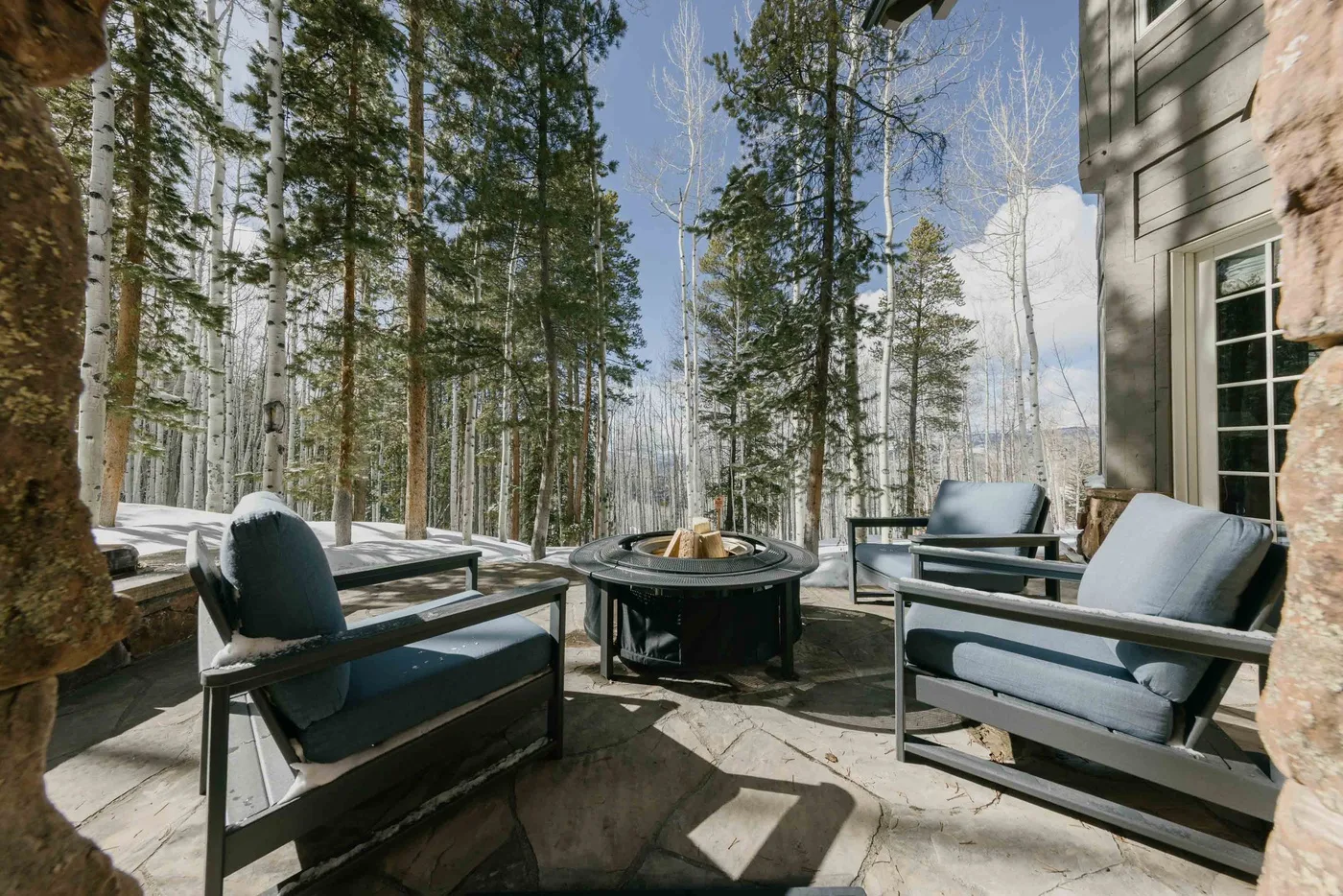 An outdoor patio with cushioned seating and a round fire pit among trees.