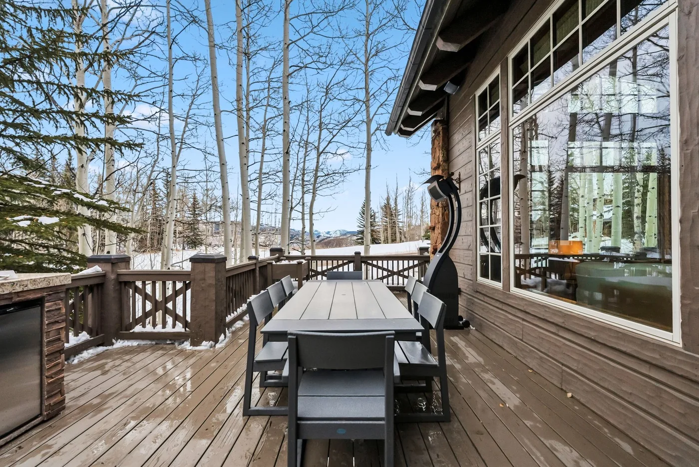 An outdoor deck with a long dining table beside large windows.