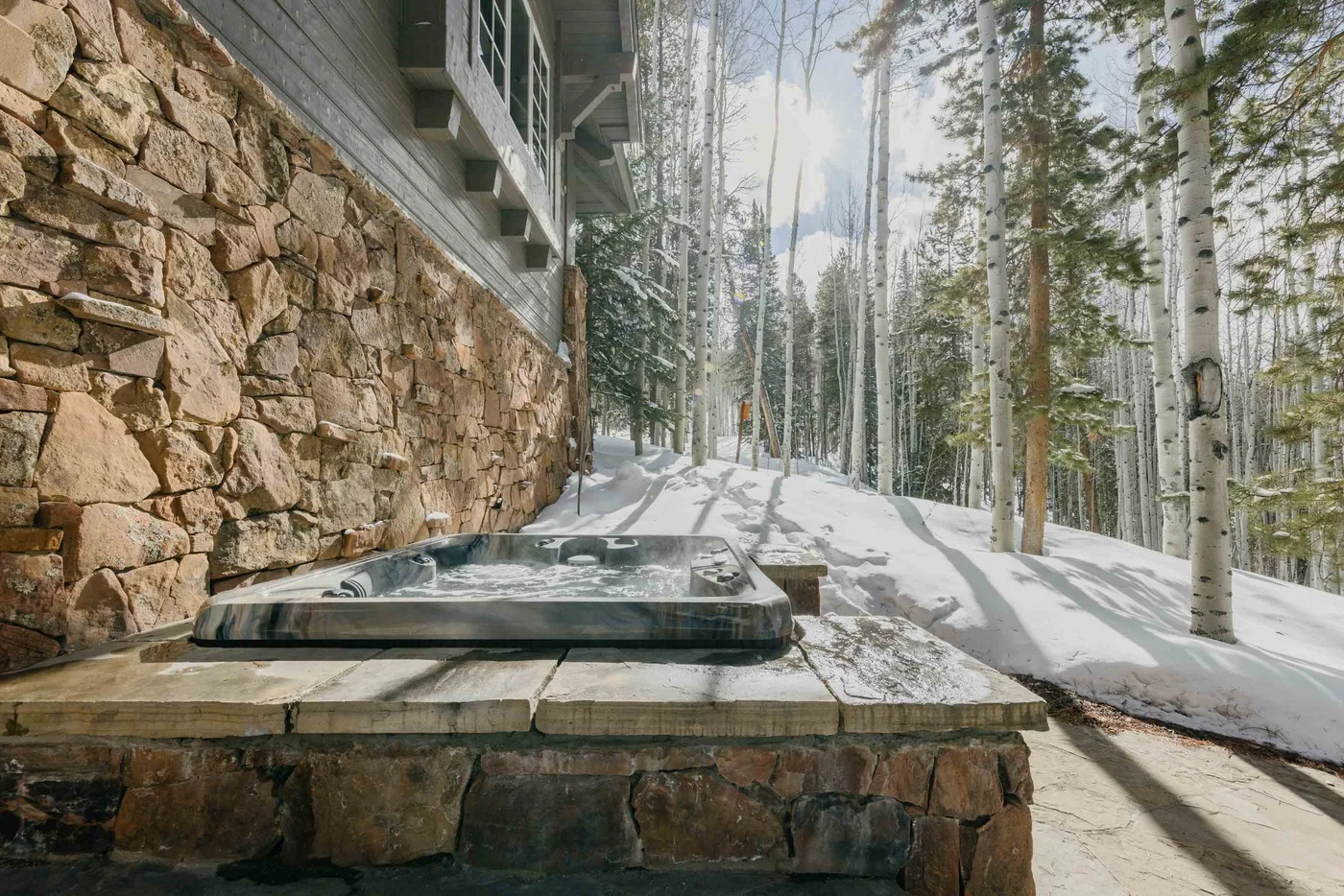 An outdoor hot tub beside a stone wall, with snowy trees beyond.