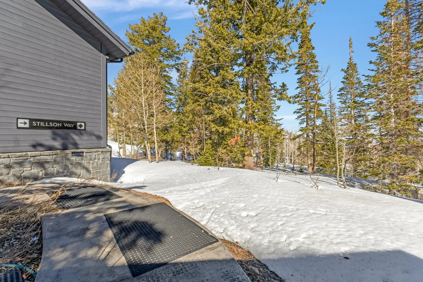 A snow-covered outdoor area beside a gray building, surrounded by evergreen trees.