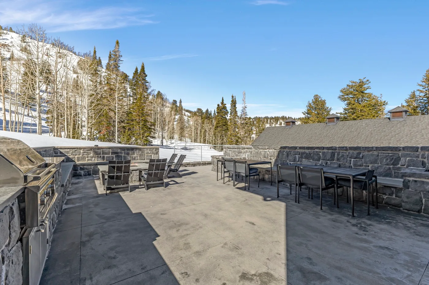 An outdoor patio with tables and seating beside a stone wall, overlooking a snowy wooded slope.