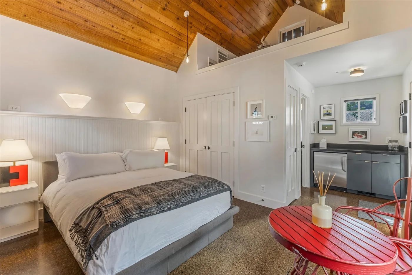 A bedroom with a bed beneath a sloped wooden ceiling and a small dining area.