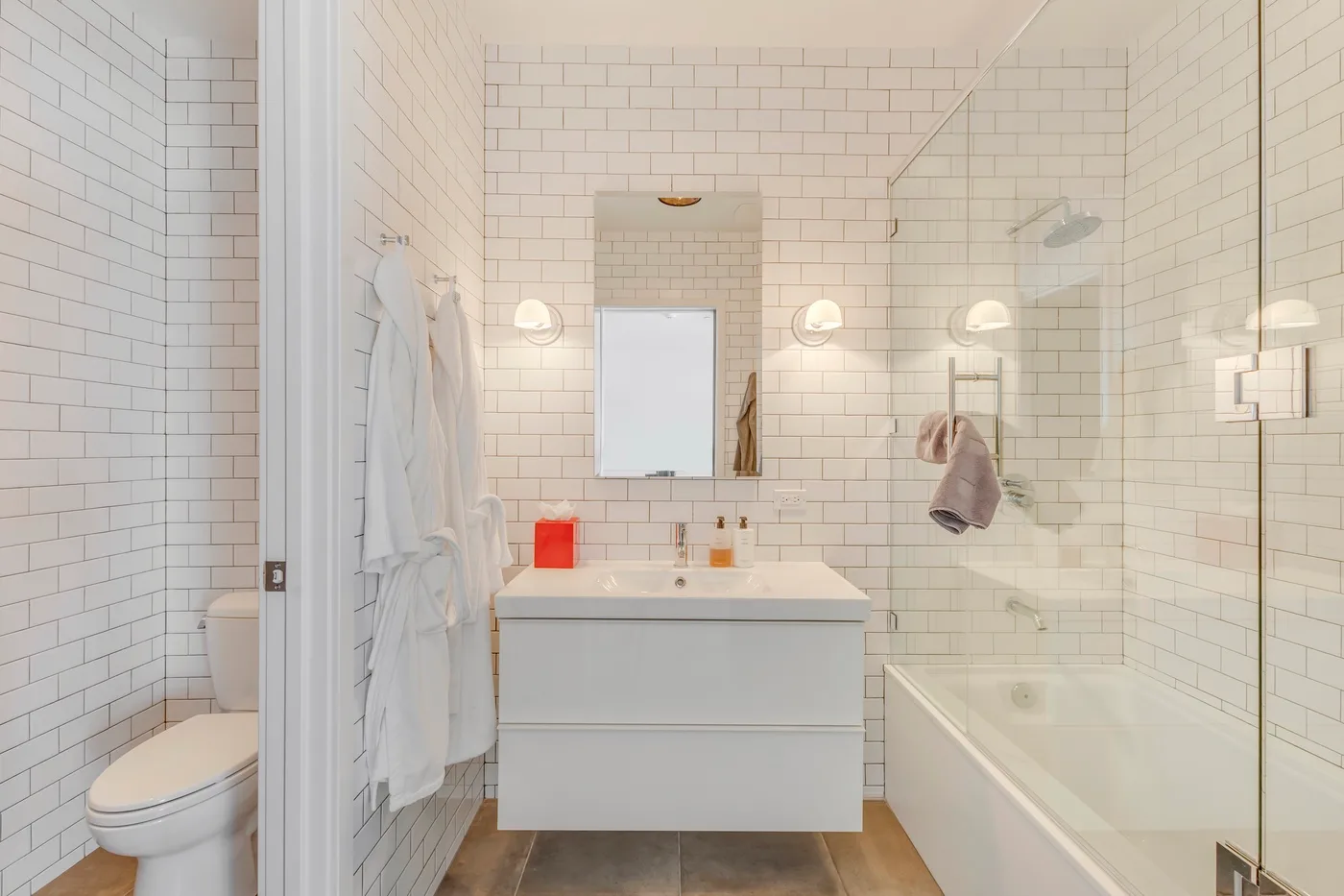 A bright tiled bathroom with a vanity, toilet, and bathtub behind a glass screen.