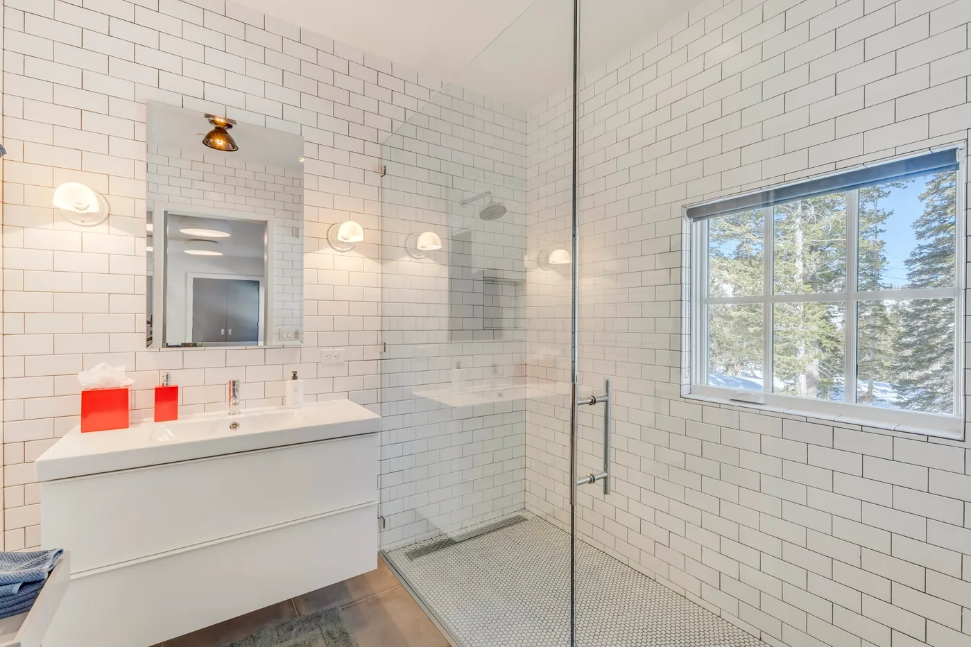 A bright tiled bathroom with a vanity, glass shower, and large window.