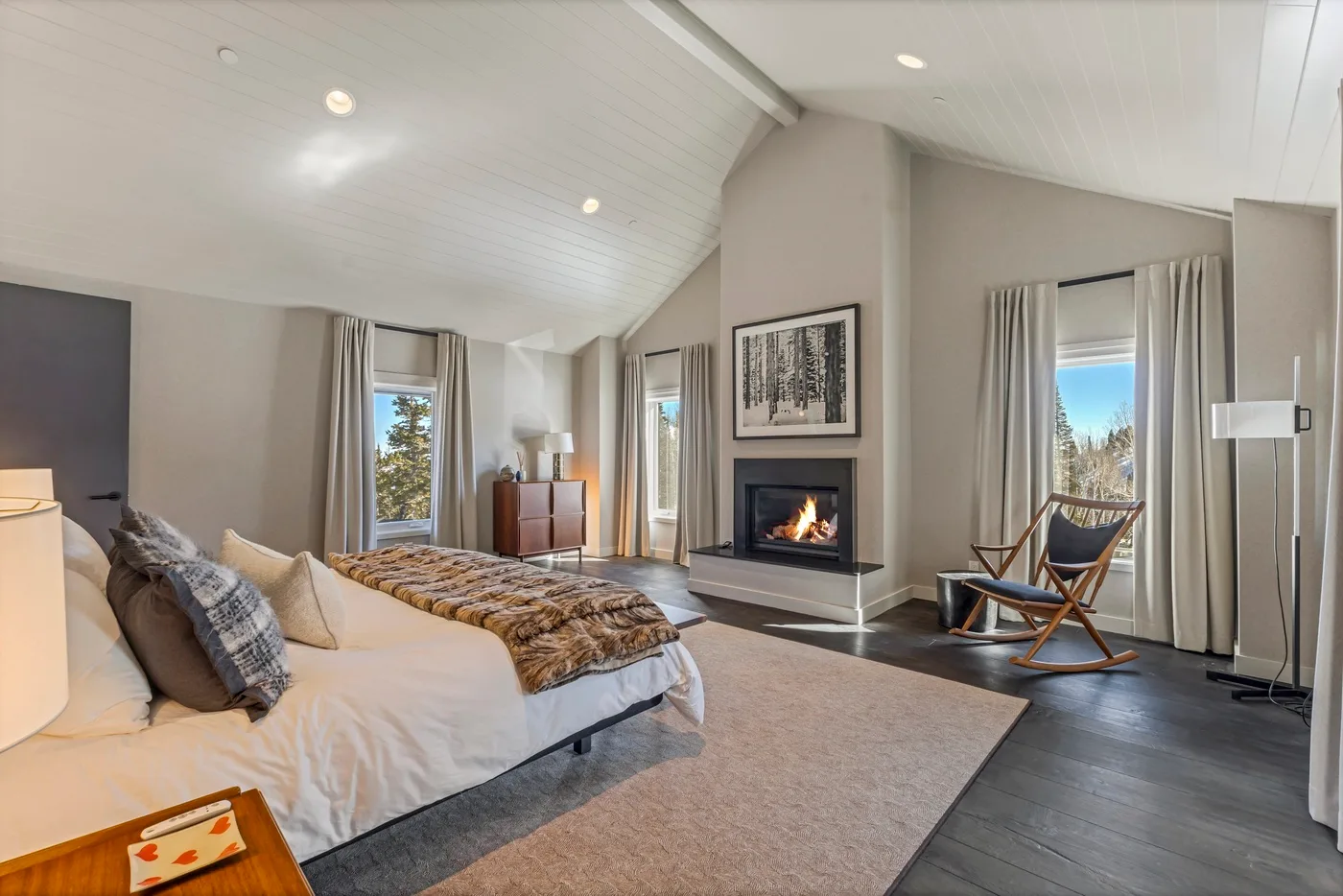 A bright bedroom features a large bed, a fireplace, and tall windows.