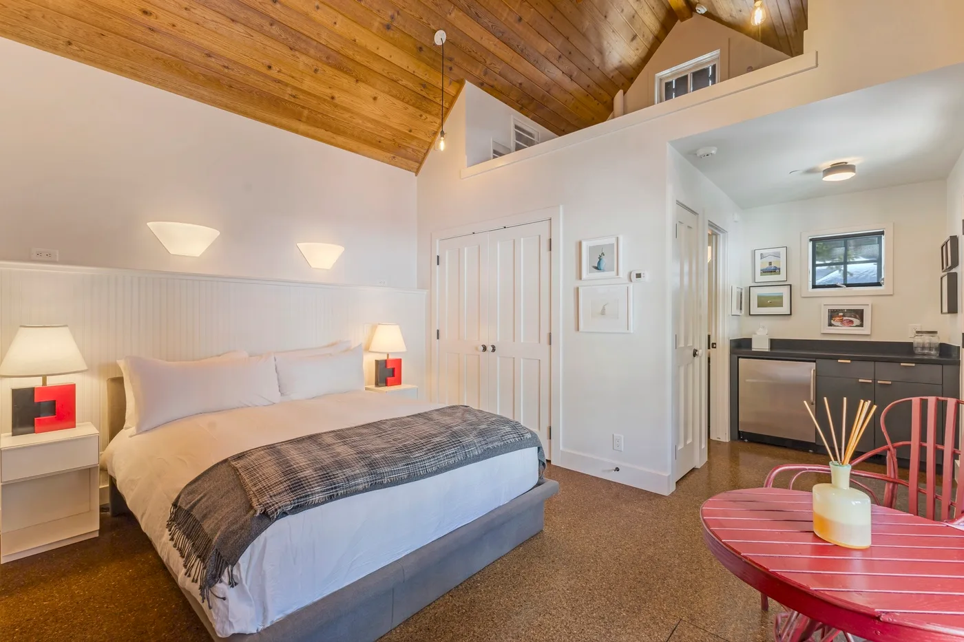 A bedroom with a bed beneath a sloped wooden ceiling and an adjoining kitchenette.